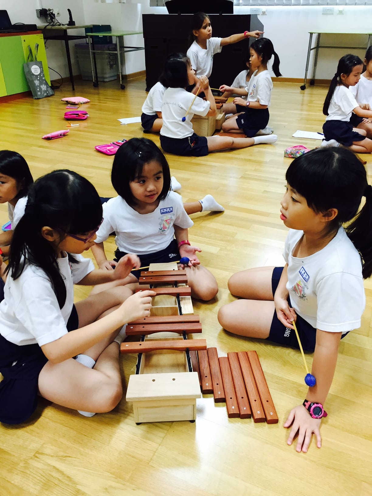 Several young girls in school uniforms sit on a wooden floor playing xylophones with mallets during a music class.
